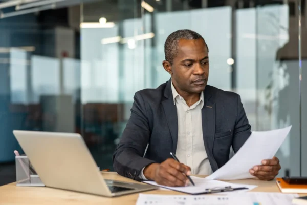 A man in business attire sits at a desk, reviewing documents with a pen in hand. A laptop and paperwork are on the desk in a modern office setting.