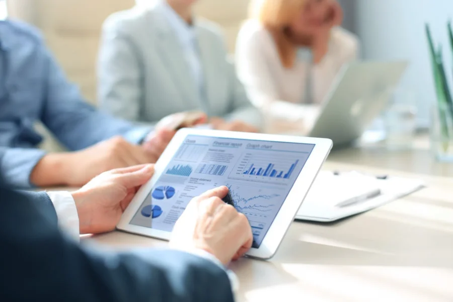 A person uses a tablet displaying charts and graphs during a business meeting with colleagues in the background.