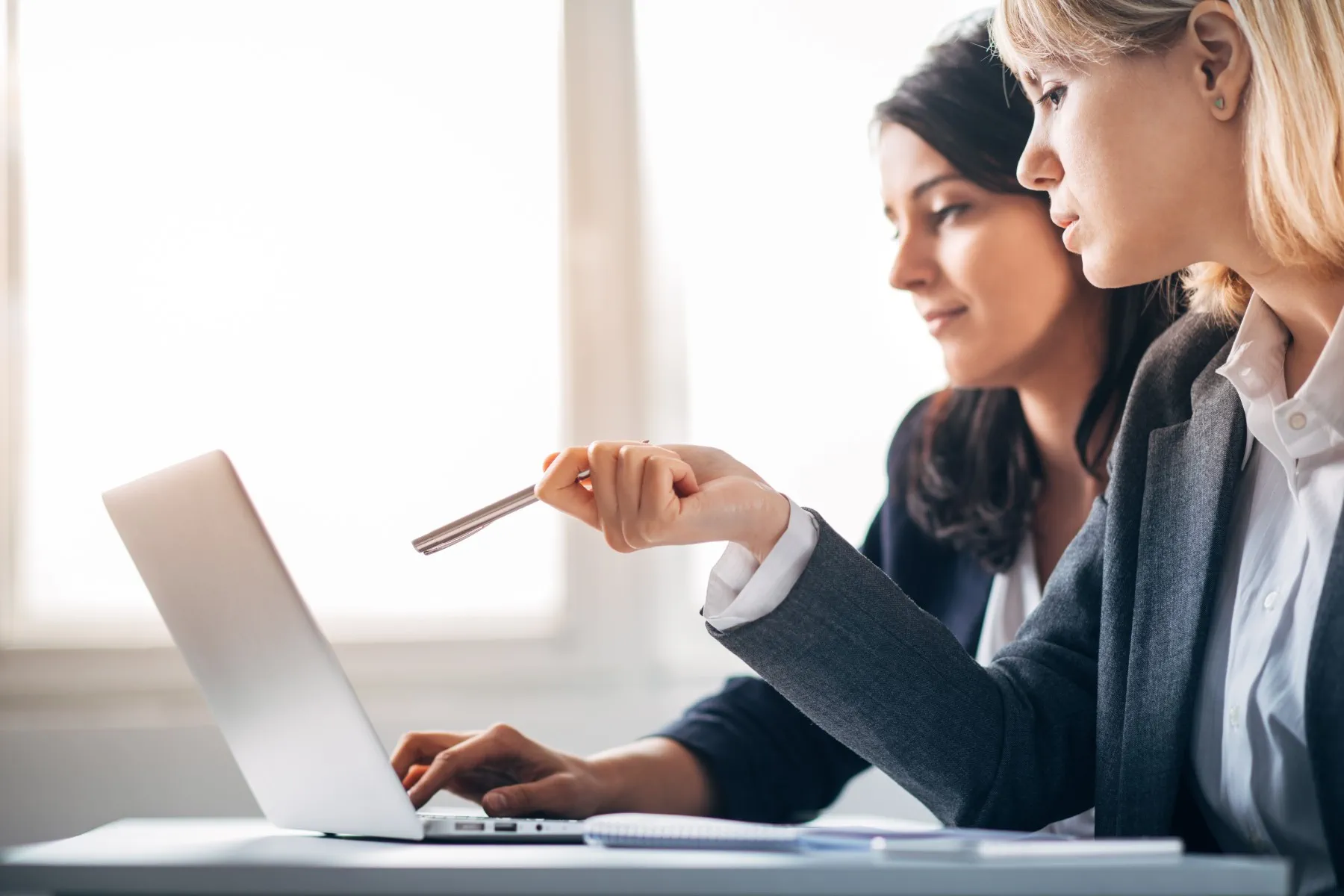 Two women in business attire work together at a laptop, with one pointing at the screen while the other types. Bright natural light comes through the window in the background.