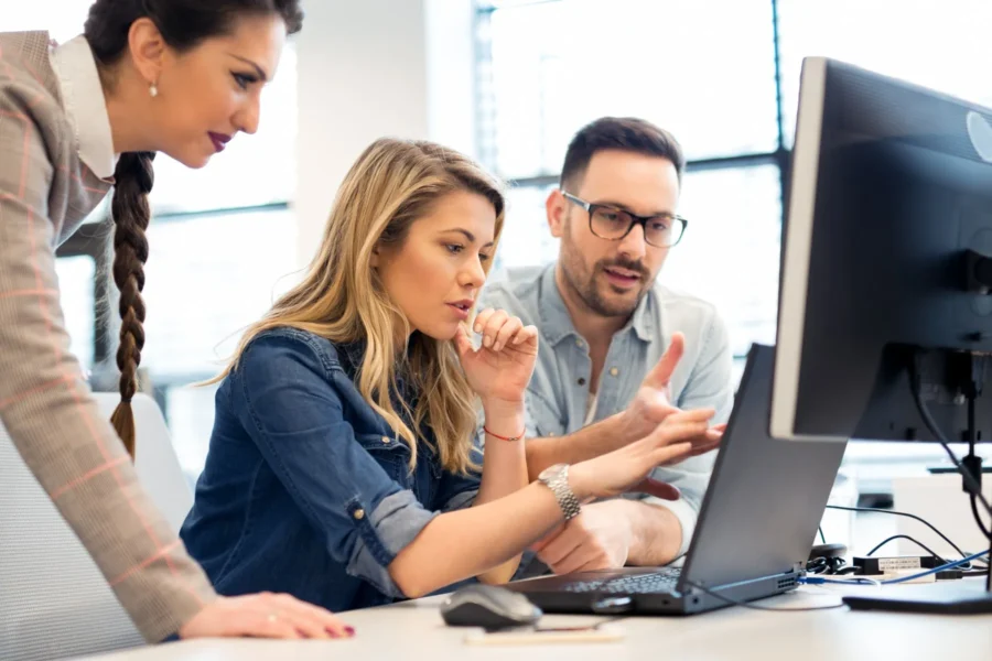 Three people sit at a desk, looking at a computer monitor together and discussing something on the screen in a bright office setting.