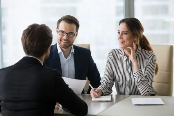 Two people sit across a table from a third person, holding papers and smiling, in a professional office setting.