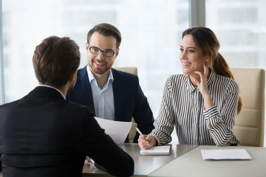 Two people sit across a table from a third person, holding papers and smiling, in a professional office setting.