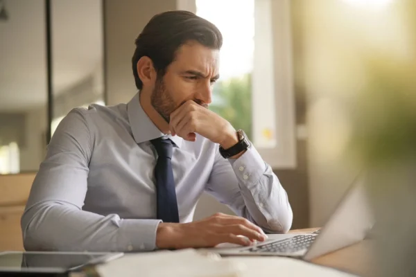 Man in a dress shirt and tie sits at a desk, looking thoughtful as he works on a laptop, with natural light coming through a window in the background.