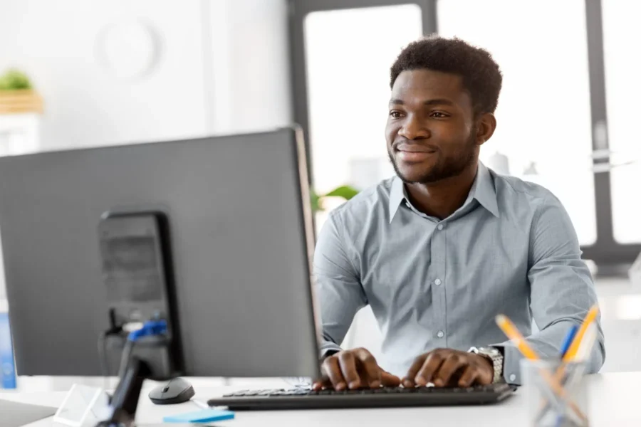 A man in a collared shirt sits at a desk, typing on a keyboard and looking at a computer monitor in a brightly lit office.