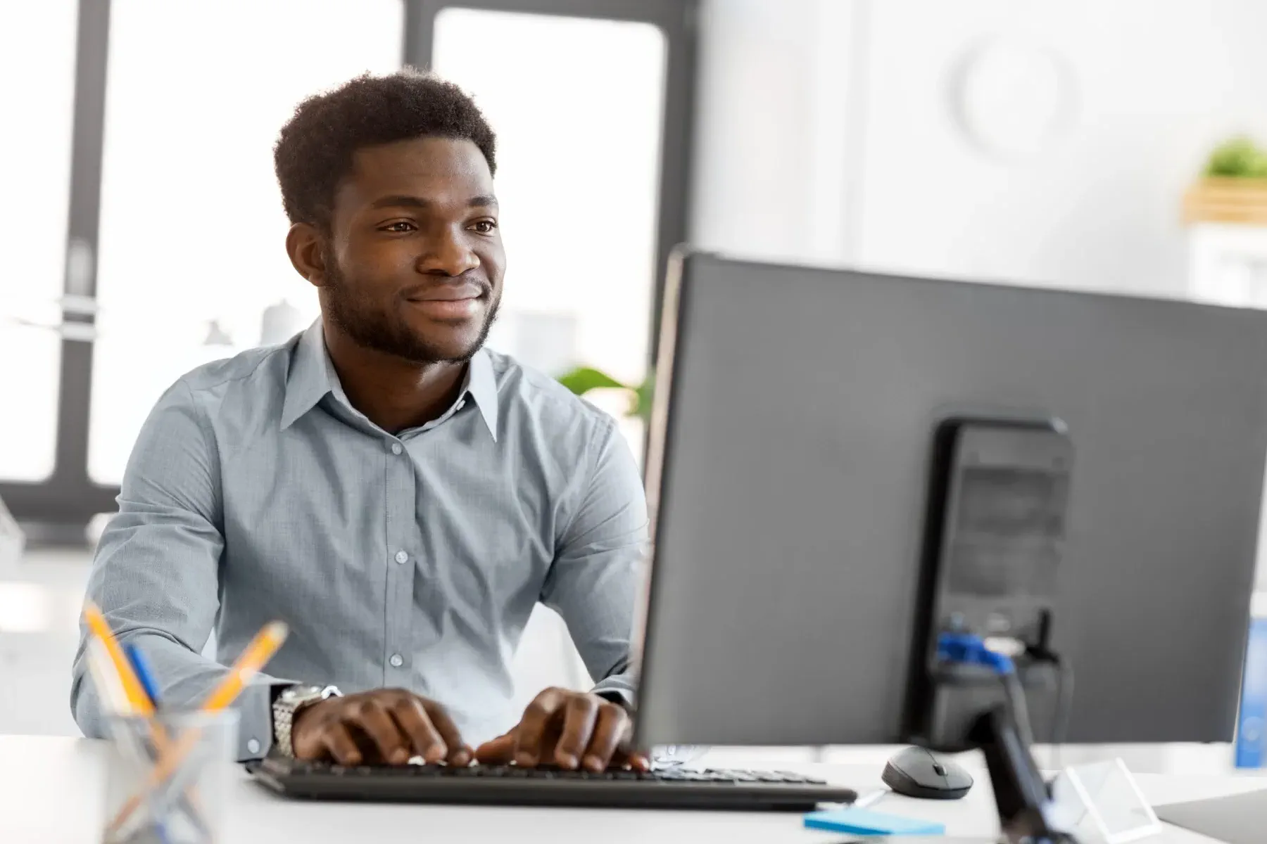 A man in a button-up shirt is working at a computer desk in a bright office, typing on a keyboard and looking at the monitor.