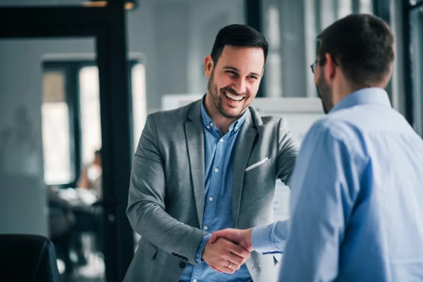 Two men in business attire are smiling and shaking hands in an office setting.