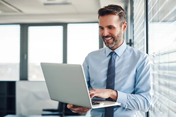 A man in a blue shirt and tie is standing by a window, smiling and typing on a laptop in a modern office setting.