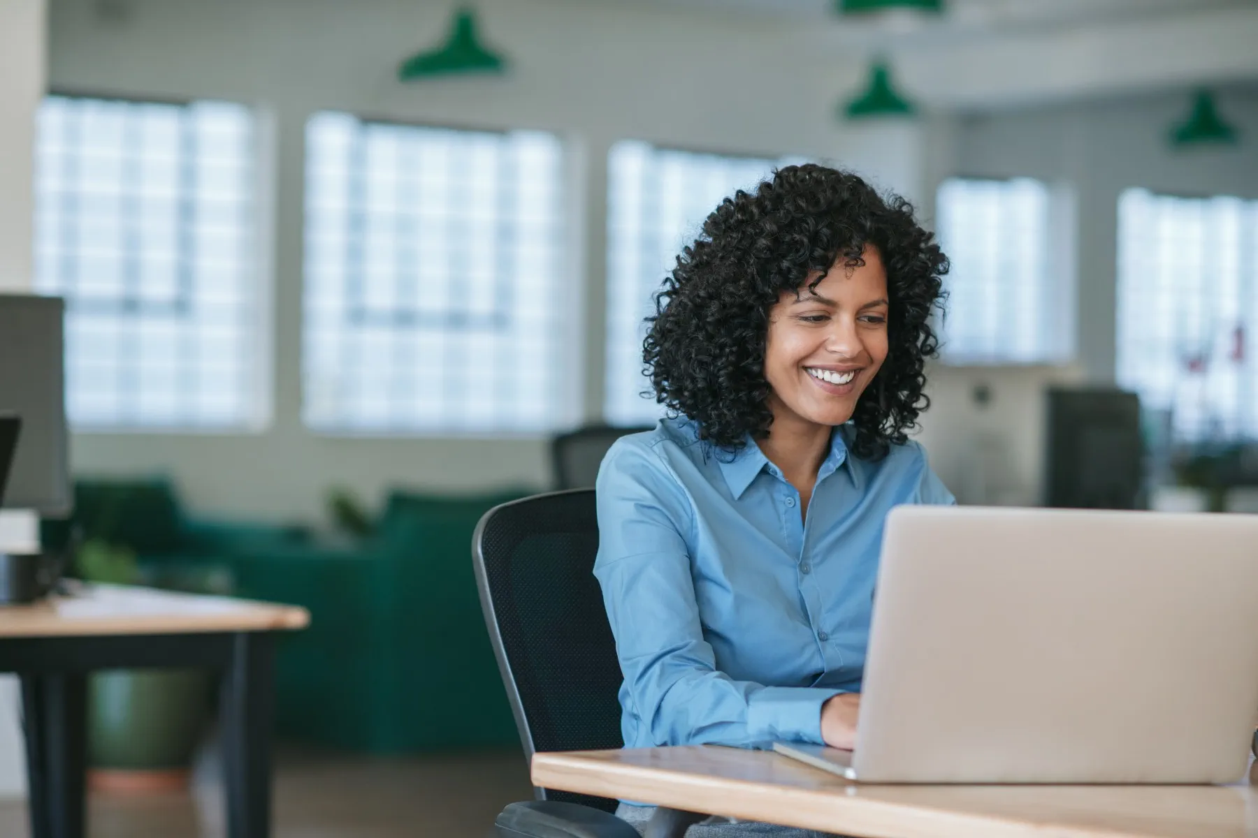 A woman with curly hair wearing a blue shirt sits at a desk and works on a laptop in a modern, bright office space.