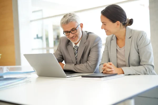 Two business professionals in suits sit at a desk, looking at a laptop screen and smiling, with documents and a notebook on the table.
