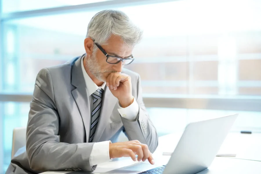 Older man in a gray suit and glasses sits at a desk, looking intently at a laptop screen while resting his chin on his hand.