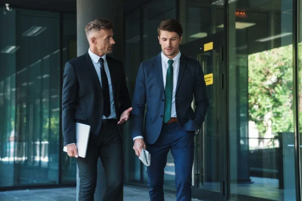 Two men in business suits walk and talk outside a glass office building; one holds a laptop, the other holds papers.