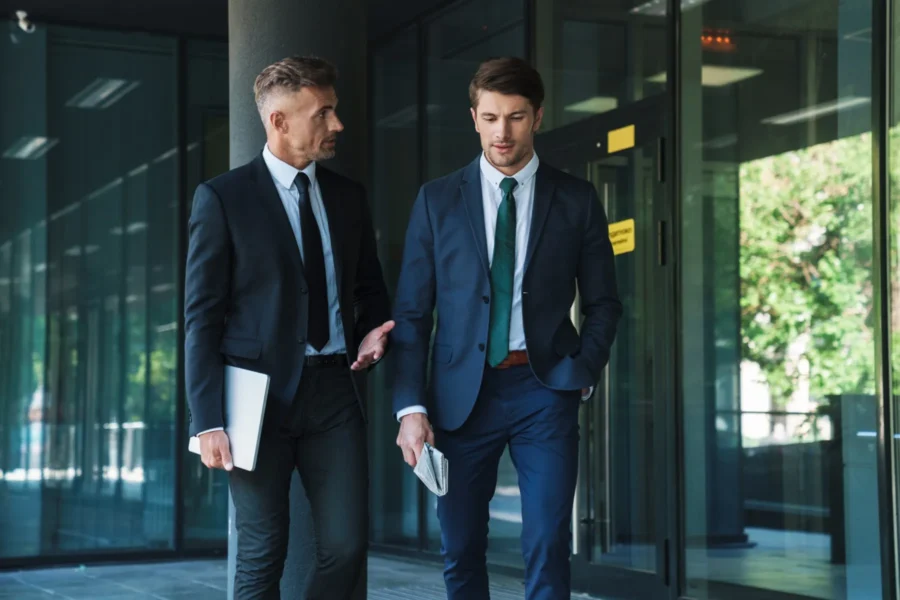 Two men in business suits walk and talk outside a glass office building; one holds a laptop, the other holds papers.