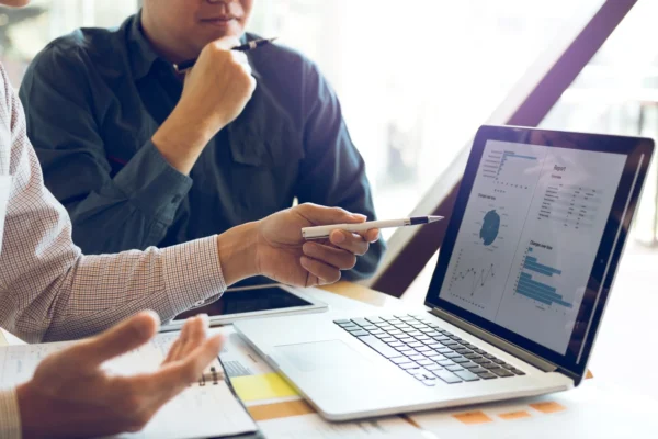 Two people sit at a desk reviewing charts and graphs on a laptop screen, with one person pointing at the display using a pen.