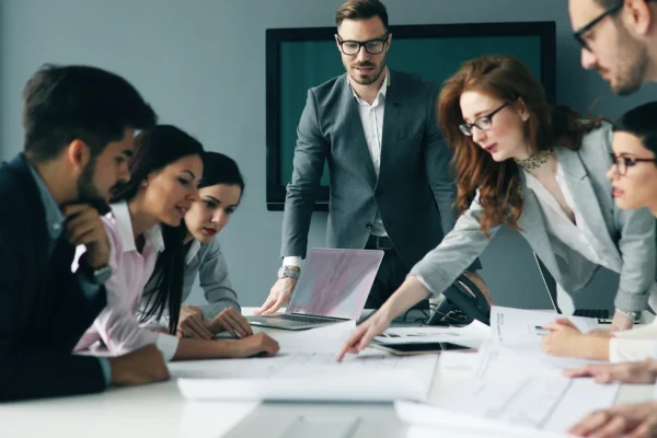 A group of business professionals in formal attire collaborate around a conference table with documents and laptops during a meeting.
