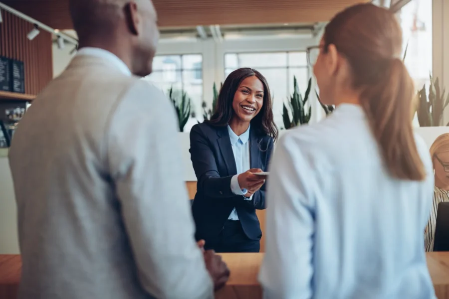 A woman in business attire hands a card to a woman across a counter while a man stands beside her in a bright office setting.