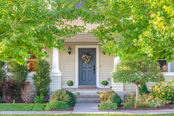 A single-story house with a gray front door, a wreath, white trim, a small porch, columns, and landscaped shrubs and trees in the front yard.