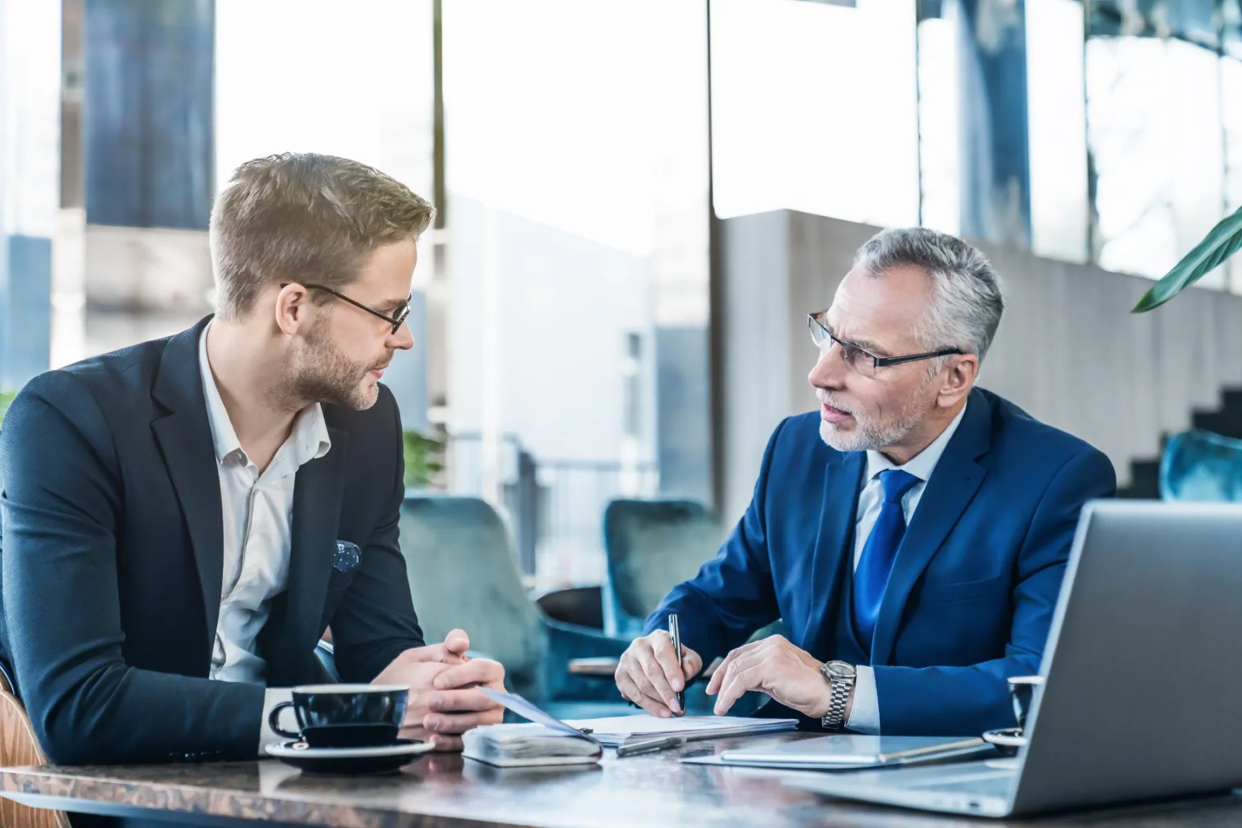 Two men in business attire sit at a table with a laptop, coffee cups, and documents, engaged in a serious conversation in a modern office setting.