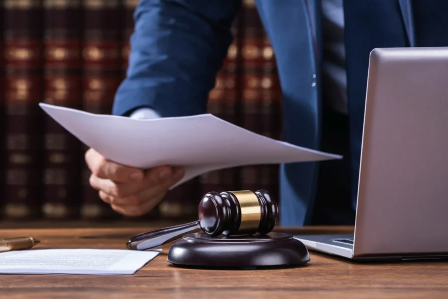 A person in a suit holding papers at a desk with a gavel, laptop, and legal books in the background.