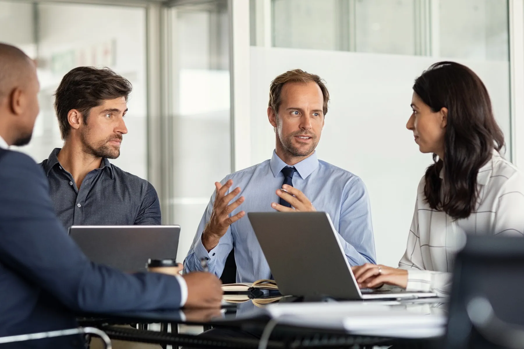 Four people sit at a table with laptops, engaged in a business meeting or discussion in a modern office setting.