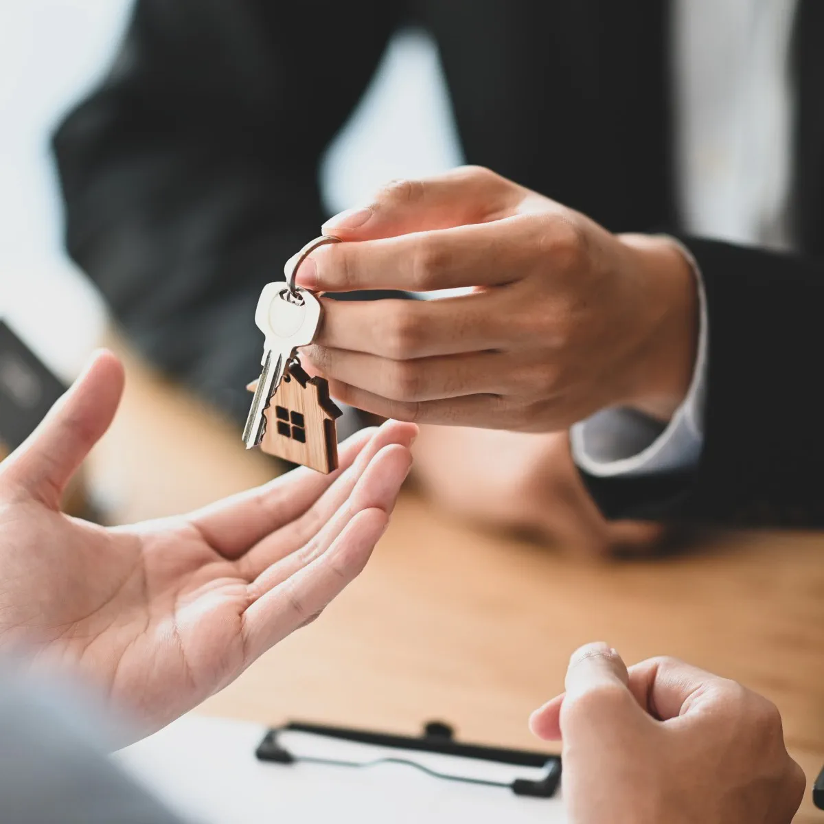 A person hands over a set of keys with a house-shaped keychain to another person, suggesting a real estate transaction.