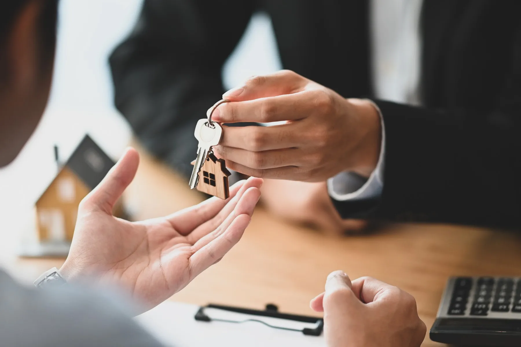 A person in a suit hands over a house-shaped keychain with keys to another person, suggesting a real estate transaction or home purchase.