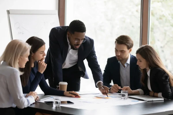 Five people in business attire sit and stand around a table, looking at documents and charts, collaborating in a modern office meeting room.