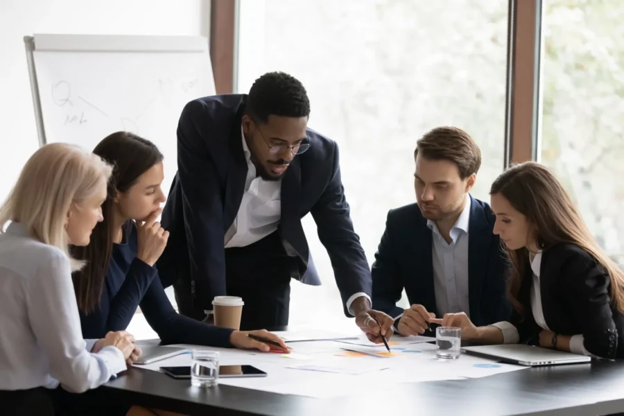Five people in business attire sit and stand around a table, looking at documents and charts, collaborating in a modern office meeting room.