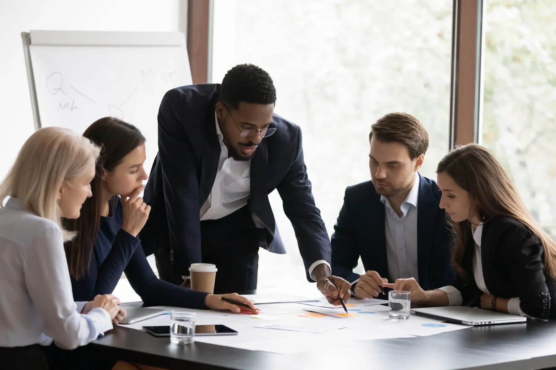 Five people in business attire sit and stand around a table, looking at documents and charts, collaborating in a modern office meeting room.