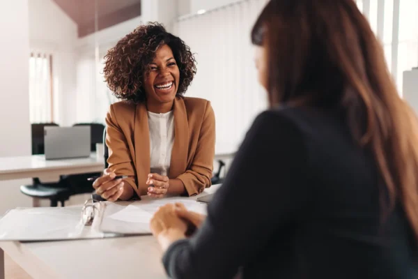 Two women sit across a desk in an office, engaged in conversation. One woman is smiling and holding a pen, while documents are visible on the desk between them.