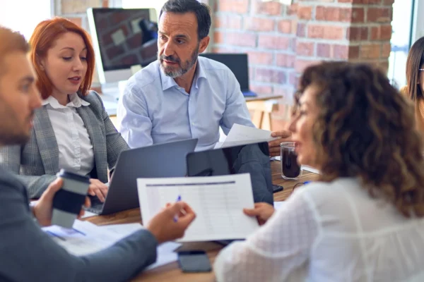 Four people sit around a table in an office, discussing documents and using laptops during a business meeting.