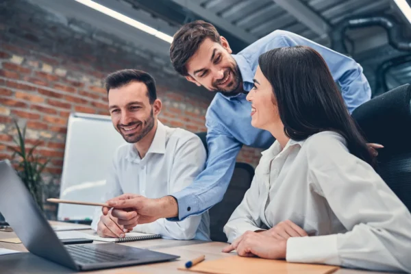 Three colleagues in business attire sit around a table, smiling and looking at a laptop during a meeting in an office with a brick wall background.