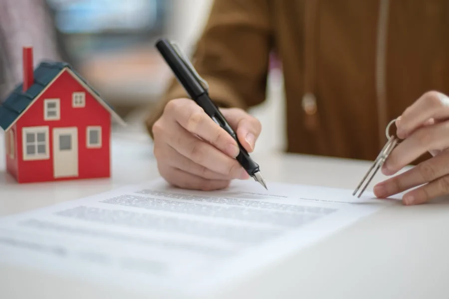 Person signing a document while holding house keys, with a small model house on the table, suggesting a real estate or home purchase transaction.