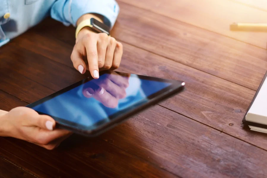 A person uses a tablet with one hand while tapping the screen with the other, seated at a wooden table next to a notebook.