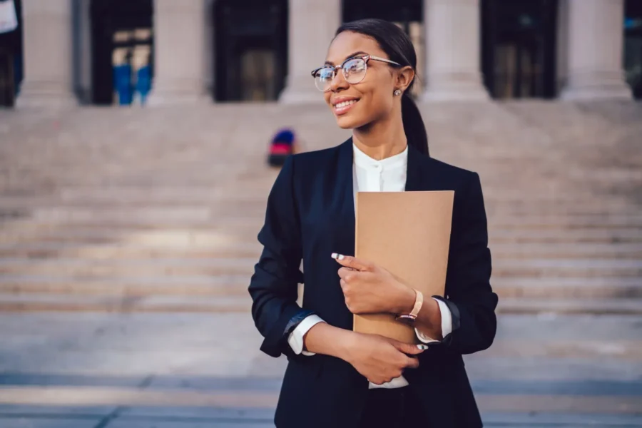 A woman wearing glasses and a black blazer holds a folder while standing outside in front of wide stone steps.