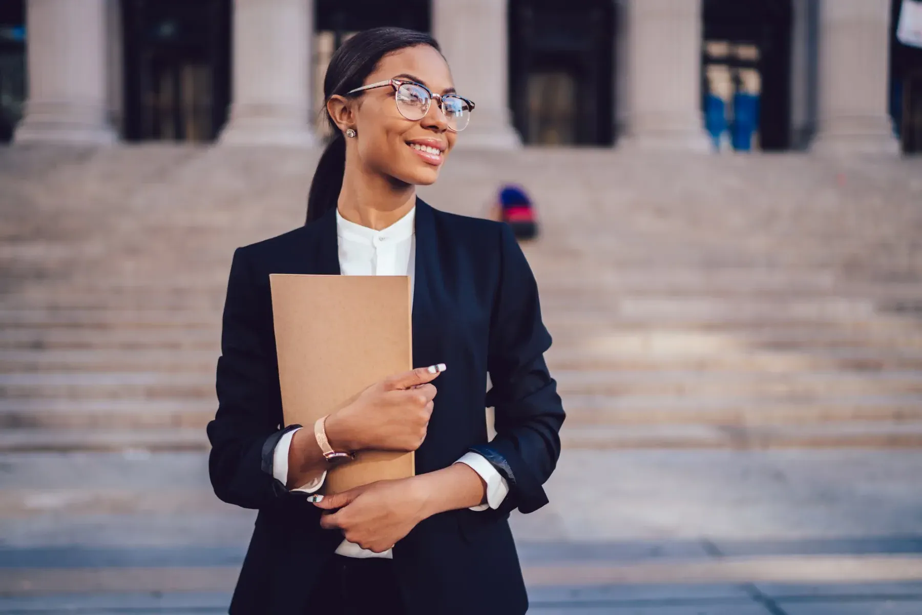A woman in business attire holds a folder and stands outside a building with large stone steps and columns in the background.