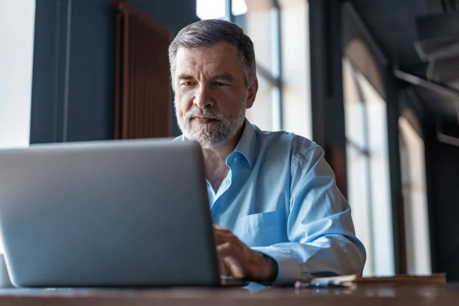Middle-aged man with gray hair and beard working on a laptop at a desk in a well-lit office or workspace.