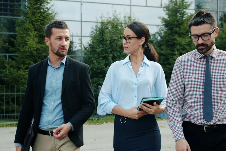 Three people in business attire walk outdoors, engaged in conversation. The woman in the center holds a tablet. Trees and a glass building are visible in the background.
