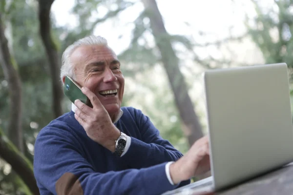 An older man smiles while talking on his cellphone and using a laptop outdoors, with trees visible in the background.