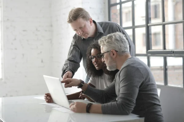 Three people sit at a desk looking at a laptop, with one person standing and pointing at the screen while the other two sit and watch attentively.