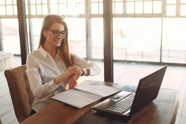 A woman in a white shirt sits at a desk with a laptop and open notebook, smiling and looking at her wristwatch in a sunlit room.