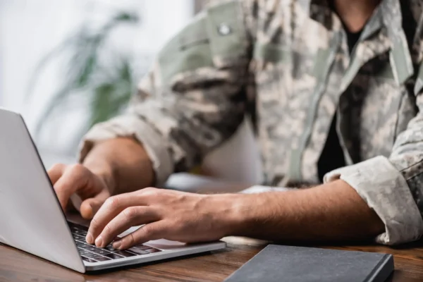 Person in camouflage uniform typing on a laptop at a desk, with a closed notebook nearby.