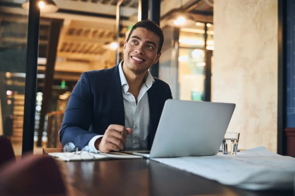 A man in a business suit sits at a desk with a laptop, notebook, glasses, and a glass of water, looking up and smiling in a modern office setting.