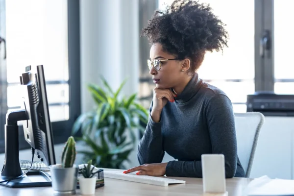 A woman wearing glasses works at a desktop computer in a modern office, surrounded by plants and office supplies.