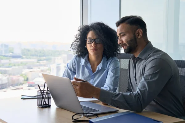 Two people sitting at a desk in an office, looking at a laptop screen and discussing something, with city buildings visible through the window behind them.