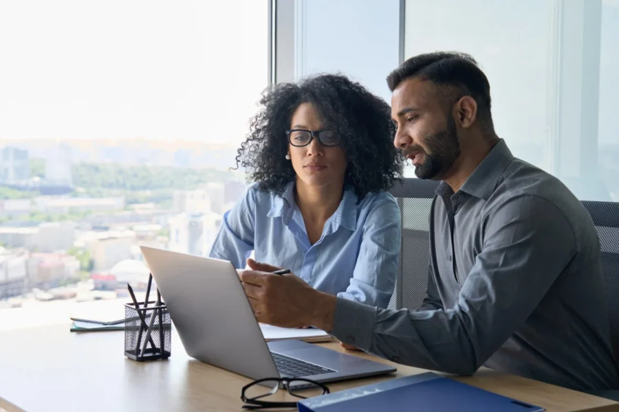Two people sitting at a desk in an office, looking at a laptop screen and discussing something, with city buildings visible through the window behind them.