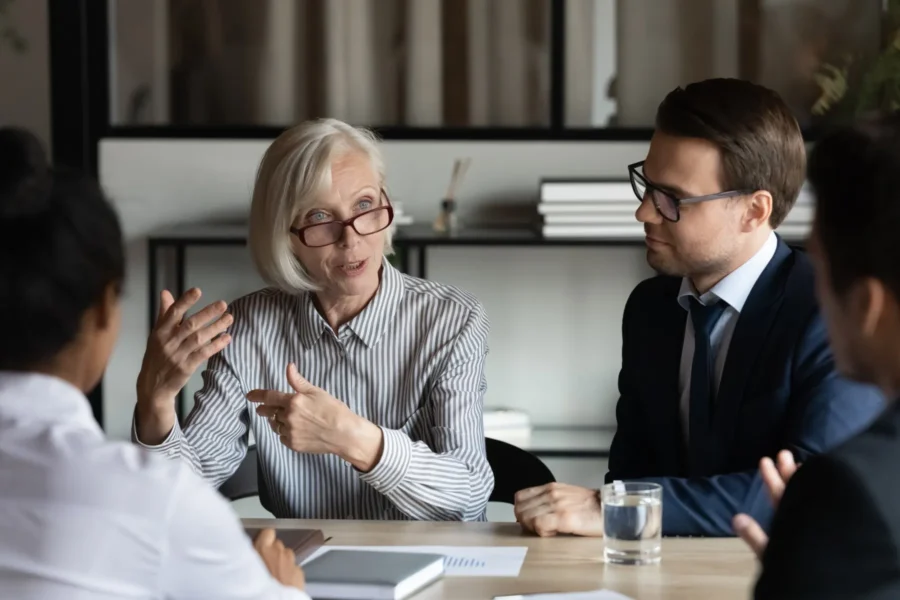 An older woman in glasses speaks while gesturing with her hands during a business meeting with three colleagues at a table.