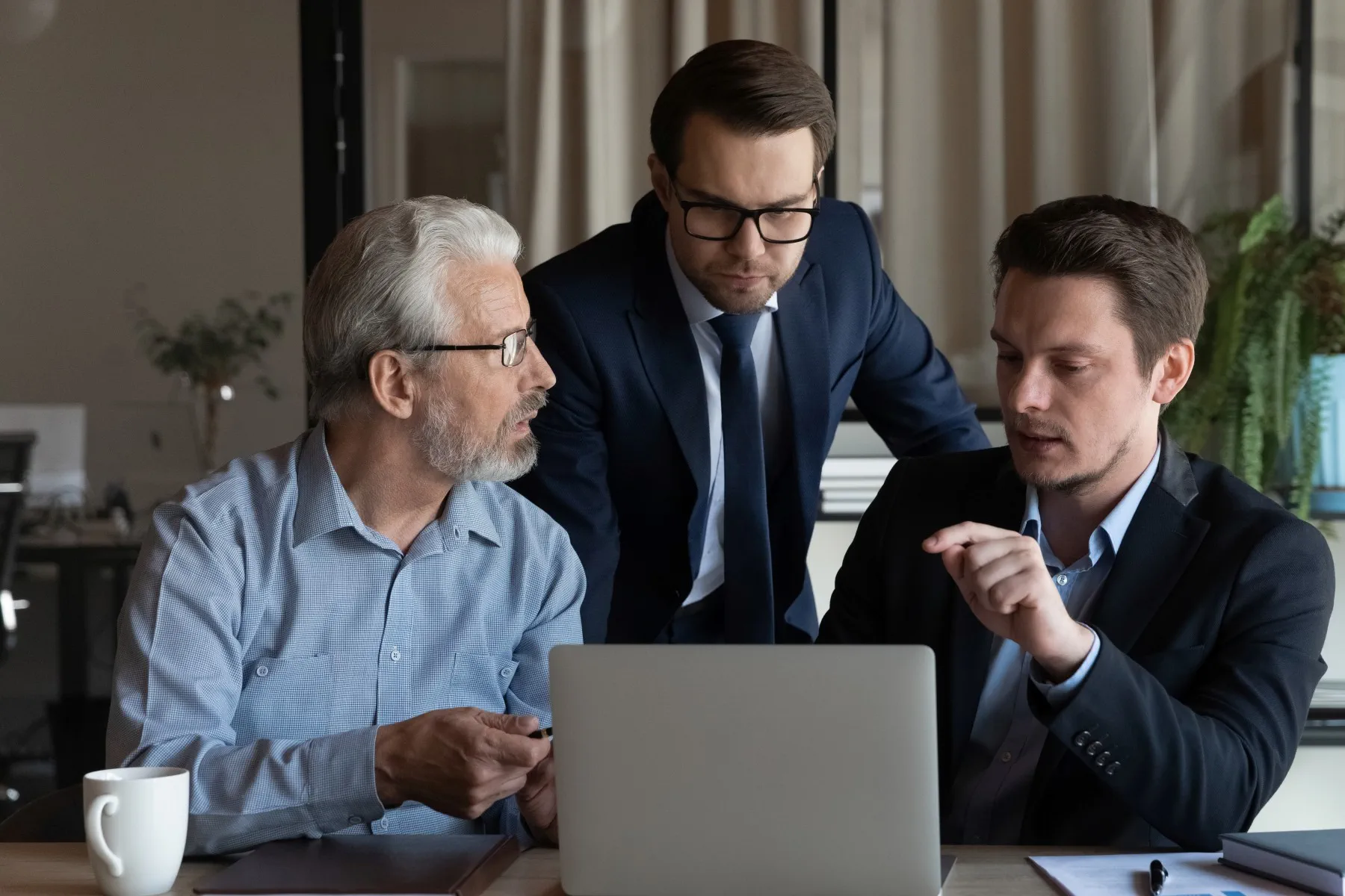 Three men in business attire are gathered around a laptop, engaged in a discussion at an office desk.