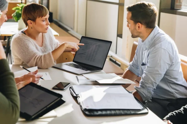 Three people sit at a table with papers, a tablet, and a laptop displaying charts, discussing data in a brightly lit office setting.