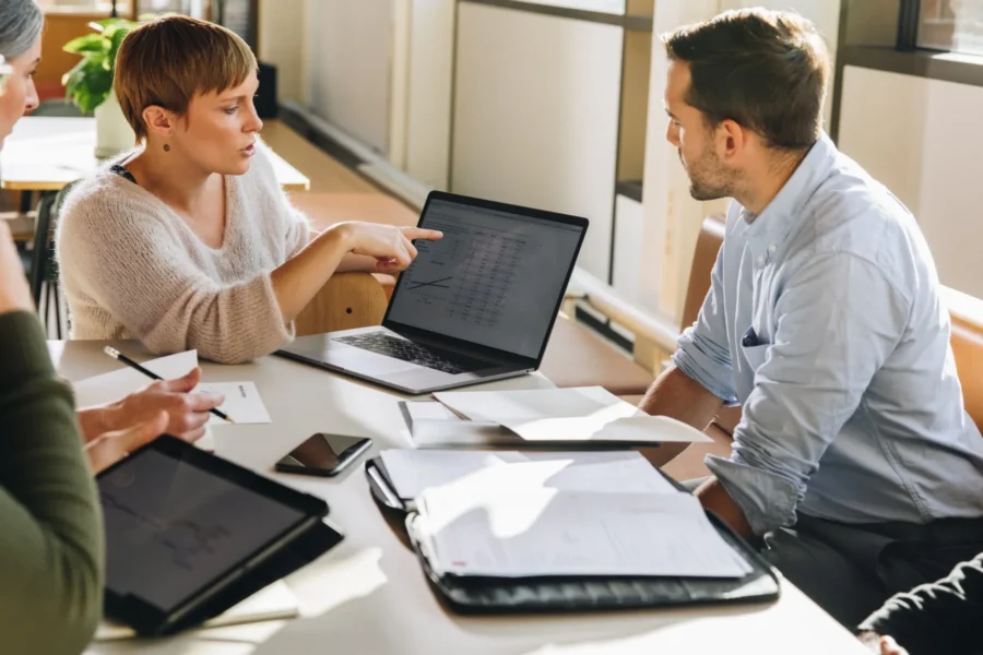 Three people sit at a table with papers, a tablet, and a laptop displaying charts, discussing data in a brightly lit office setting.