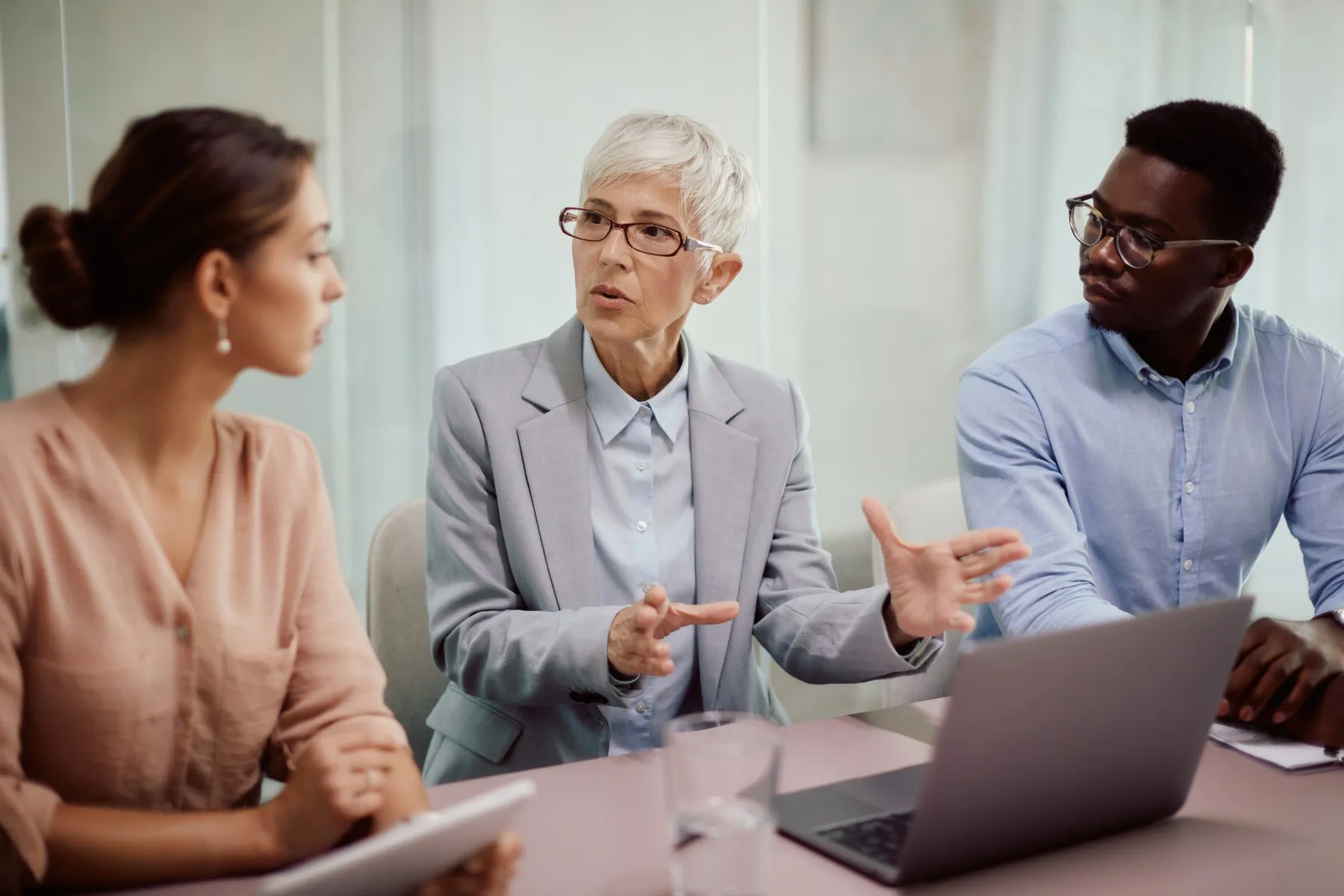Three people in business attire sit at a table with a laptop, engaged in a discussion. The woman in the middle gestures while speaking to her colleagues.